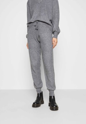Personne portant un pantalon de jogging gris en maille avec taille à cordon et des bottines noires, debout devant un fond clair uni.