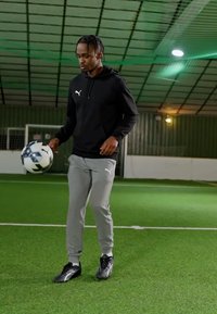 Melnā kapučjaka, pelēkas sporta bikses un melni futbola apavi. Spēlētājs līdzsvaro melnbaltu futbola bumbu uz zaļā mākslīgā zālāja.
