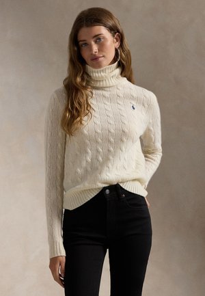 Žena s dlhými vlnitými vlasmi, oblečená v krémovom pletenom roláku s vrkočovým vzorom a čiernych nohaviciach, stojaca pred jednoliatym béžovým pozadím.