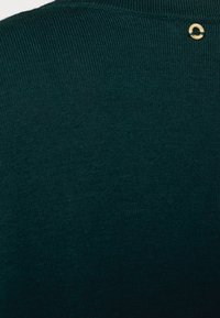 Tejido de suéter de punto de color verde oscuro con una textura suave, que presenta un pequeño anillo de oro como acento cerca del escote.