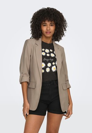 Donna con capelli ricci che indossa un blazer beige, pantaloncini neri e una T-shirt nera con grafiche di margherite bianche e la scritta "Daisy days".