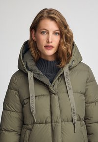 Olivgrön puffjacka med huva, med zip-för closure, kviltat mönster och snoddar med logotyp. Tjockt, isolerat tyg.