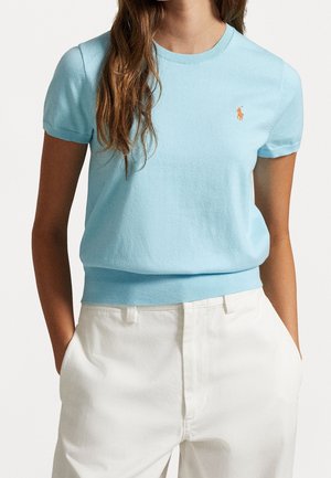 Vrouw draagt een lichtblauwe gebreide top met korte mouwen en een klein oranje logo en witte high-waist broek met de handen in de zakken.
