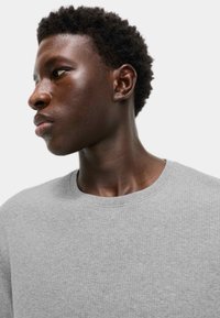 Young Black man with short curly hair wearing a light gray ribbed crew neck shirt, looking to the left on a plain white background.