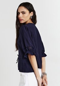 Donna con lunghi capelli scuri, che indossa una maglietta blu navy con maniche a sbuffo e pantaloni bianchi, rivolta di lato, con orecchini e braccialetti d'argento.