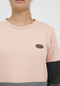 Sweatshirt en coton rose clair avec des accents gris. Doté d'un col rond et d'un patch oval en logo marron sur la poitrine. Texture douce dans l'ensemble.