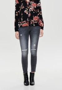 Mujer vestida con jeans skinny negros descoloridos con parches desgastados, camisa de botones negra con estampado floral, y botas negras hasta el tobillo sobre un fondo plano.