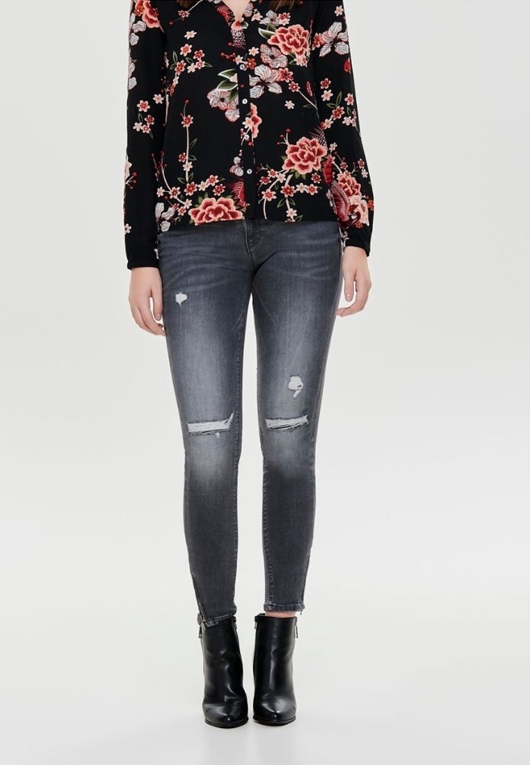 Mujer vestida con jeans skinny negros descoloridos con parches desgastados, camisa de botones negra con estampado floral, y botas negras hasta el tobillo sobre un fondo plano.