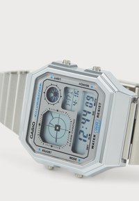 Montre numérique argentée en forme rectangulaire, avec un affichage bleu clair, plusieurs boutons et un bracelet en acier inoxydable. Résistante à l'eau.