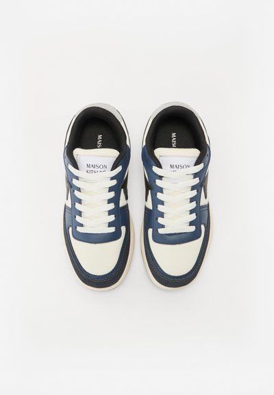 Zapatillas marinas y crema con parte superior de cuero suave, cordones blancos y un interior negro. Presenta una etiqueta de logo en la lengüeta y acentos texturizados en contraste.
