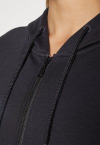 Hoodie noir à fermeture éclair avec cordons de serrage porté, fermeture éclair partiellement fermée au niveau du cou, dévoilant le cou et le haut de la poitrine de la personne.