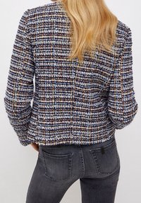 Giacca in tweed blu, marrone e crema; motivo a quadri texturizzato; design aderente con maniche lunghe; vista posteriore; abbinata a denim scuro.