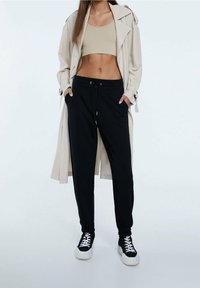 Frau steht mit Händen in den schwarzen Jogginghosen-Taschen, trägt ein beige Crop-Top, einen beige Trenchcoat und schwarze Sneaker mit weißen Sohlen.