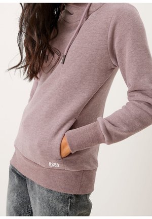Vrouw draagt een mauve hoodie met hand in de zak en vervaagde zwarte jeans, afgebeeld van nek tot heupen tegen een effen achtergrond.
