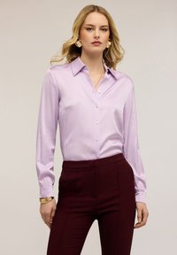 Kvinde iført en lys lilla satin skjorte med knapper, mørkerøde højtaljede bukser, guldringe og armbånd på en neutral baggrund.