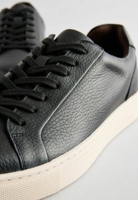 Adidași negri din piele cu o suprafață texturată, șireturi negre și o talpă din cauciuc alb. Prezintă o cusătură subtilă și un design minimalist.