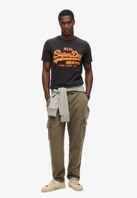 Camiseta de algodón negra con logotipo "SuperDry" naranja, sudadera gris atada a la cintura, pantalones cargo caqui, zapatillas de deporte crema.