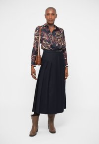 Lang marineblå nederdel med plisseringer og bæltet talje, parret med en bluse med flerfarvet mønster. Brune ankelstøvler og en lille brun taske.