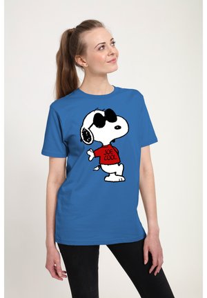 Donna con camicia blu con il personaggio Snoopy con occhiali da sole e maglione rosso "Joe Cool", in piedi davanti a uno sfondo semplice.