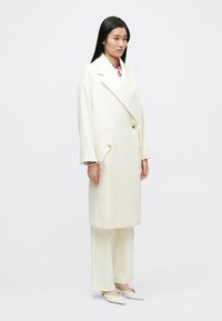 Kermanvärinen kaksirivinen takki, jossa suuret kauluskäänteet, kaksi etutaskua ja kirkkaat napit, yhdistetty samansävyisiin housuihin ja teräväkärkisiin tasapohjiin kenkiin.