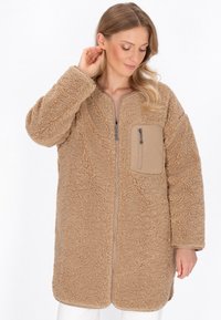 Giacca in pile beige con una superficie morbida e testurizzata, frontale con zip e taschino sul petto. Il design presenta una vestibilità rilassata e maniche lunghe.
