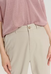 Persona che indossa pantaloni beige chiaro con bottone e passanti per cintura, abbinati a un top a maniche corte rosa in maglia infilato dentro.