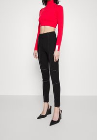 Vrouw in een rode cropped coltrui, zwarte hoge taille broek met witte stiksels, en zwarte puntige sleehakken, staande op een effen achtergrond.