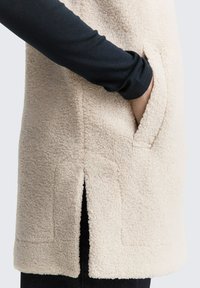 Cremfarbener Fleece-Weste mit einer Seitentasche, gespaltenem Saum und strukturiertem Stoff. Schwarzes Langarm-Oberteil ist darunter sichtbar.