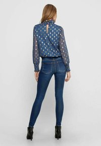 Helles blau gemustertes Oberteil aus transparentem Stoff, mit langen Ärmeln und einem Schlüsselloch-Detail im Rückenteil, kombiniert mit dunkelblauen, eng sitzenden Jeans und schwarzen High Heels.
