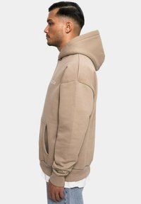 Beige Kapuzenpullover mit einer vorderen Tasche, gerippten Bündchen und Saum. Verfügt über ein einfaches Textdesign auf der Brust. Weiche Stofftextur.