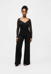 Femme debout, face à l'avant, portant une combinaison noire à manches longues transparentes et des talons ouverts noirs, sur fond blanc.