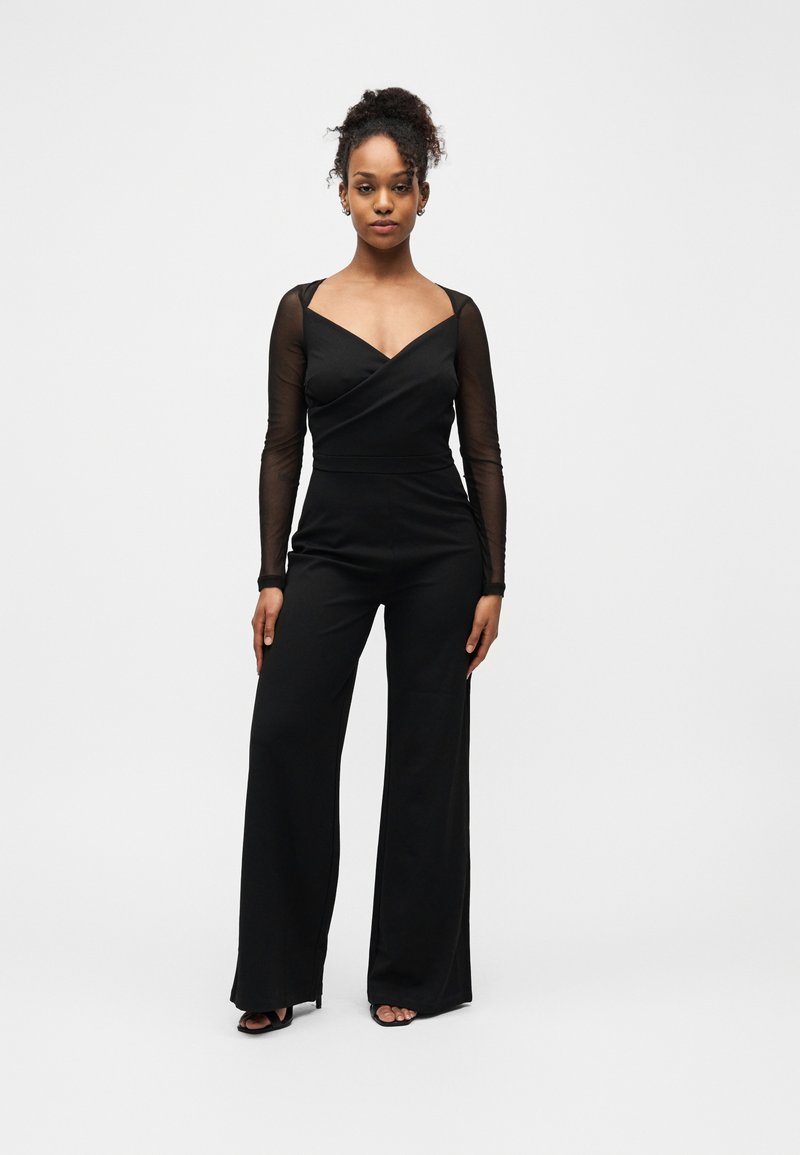 Femme debout, face à l'avant, portant une combinaison noire à manches longues transparentes et des talons ouverts noirs, sur fond blanc.