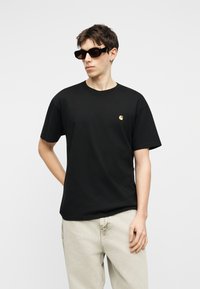 Młody mężczyzna w czarnej koszulce z logo, jasnobeżowych spodniach, ciemnych prostokątnych okularach przeciwsłonecznych, stojący z jedną ręką za plecami na jednolitym tle.