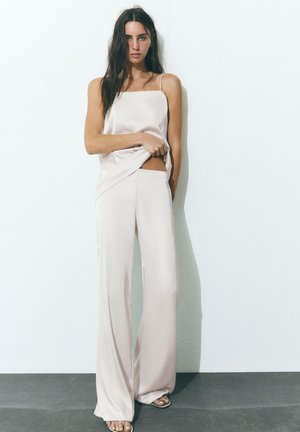 Frau mit langen dunklen Haaren, die ein ärmelloses cremefarbenes Satin-Top und eine Hose mit weitem Bein trägt, steht vor einer schlichten weißen Wand.