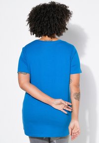 Camiseta de algodón azul con mangas cortas, cuello redondo y un corte holgado, vista desde la parte de atrás. Tatuajes visibles en los brazos.