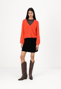 Cardigan en tricot rouge avec un décolleté en V et une fermeture par boutons, associé à une jupe en velours noir et des bottes de cowboy marron, ornées de motifs en relief.