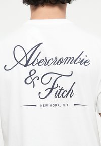 Weißes Baumwoll-T-Shirt mit auffälligem navy-farbenen Druck von "Abercrombie & Fitch" und "New York, N.Y." zentral auf dem Rücken. Runder Halsausschnitt, kurze Ärmel.