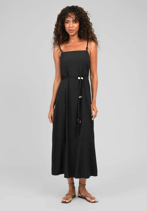 Femme portant une robe midi noire sans manches à fines bretelles, une taille ceinturée avec des perles et des sandales plates à brides, debout devant un fond uni.