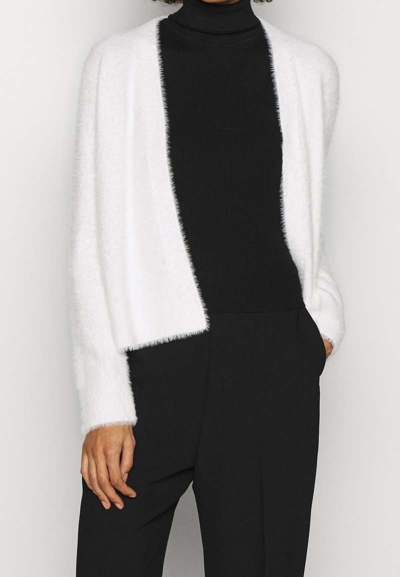 Personne portant un cardigan blanc duveteux ouvert sur un pull à col roulé noir et un pantalon noir, avec une main dans la poche.