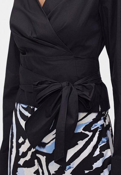 Blouse portefeuille noire à manches longues nouée en un grand nœud à la taille, associée à une jupe à motif abstrait noir, blanc et bleu.
