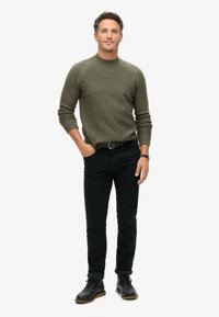 Dunkelgrüner Strickpullover mit Rundhalsausschnitt, kombiniert mit schwarzen Slim-Fit-Hosen und schwarzen Schnürschuhen, mit hochgerollten Bündchen.