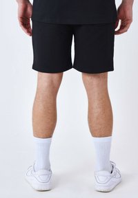 Shorts noirs en coton, avec un design simple et des bords ourlés. Portés avec des chaussettes blanches et des chaussures de sport blanches.