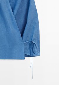 Blå linne-wraptop med lång ärm knuten vid mudd och djup V-ringning fram mot vit bakgrund.