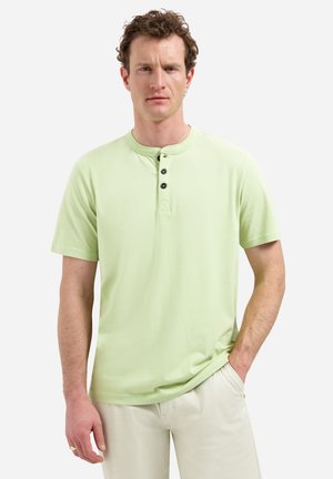 Man met krullend haar die een lichtgroen henley-shirt met korte mouwen en drie knopen draagt, staand met één hand in zijn zak.
