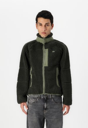 Chaqueta de forro polar en verde oscuro con una parte superior de tela lisa. Tiene dos bolsillos frontales con cremallera y un cuello alto. Acabado suave y texturizado.