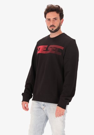 Giovane uomo con capelli ricci corti e barba, indossa una felpa nera con testo grafico rosso e jeans azzurri chiari, in piedi davanti a uno sfondo neutro.
