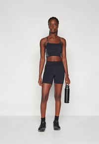 Svart sports-BH med mønstrede stropper, matchende svarte shorts og svarte joggesko. Holder en svart vannflaske. Ensfarget bakgrunn.