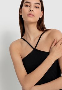 Melnā ribota topiņa ar pieguļošu dizainu. Iezīmējas ar plānām siksniņām, kas krustojas aizmugurē. Gluda tekstūra ar minimālu apdari.