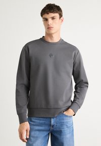 Gray sweatshirt made of soft fabric, featuring a round neckline and hexagonal logo on the chest. Styled with blue jeans.