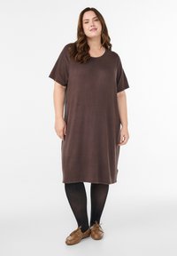 Sieviete stāv, valkājot vaļīgu brūnu kleitu ar īsām piedurknēm, melnas zeķubikses un brūnas kurpes ar šņorēm uz vienkārša balta fona.